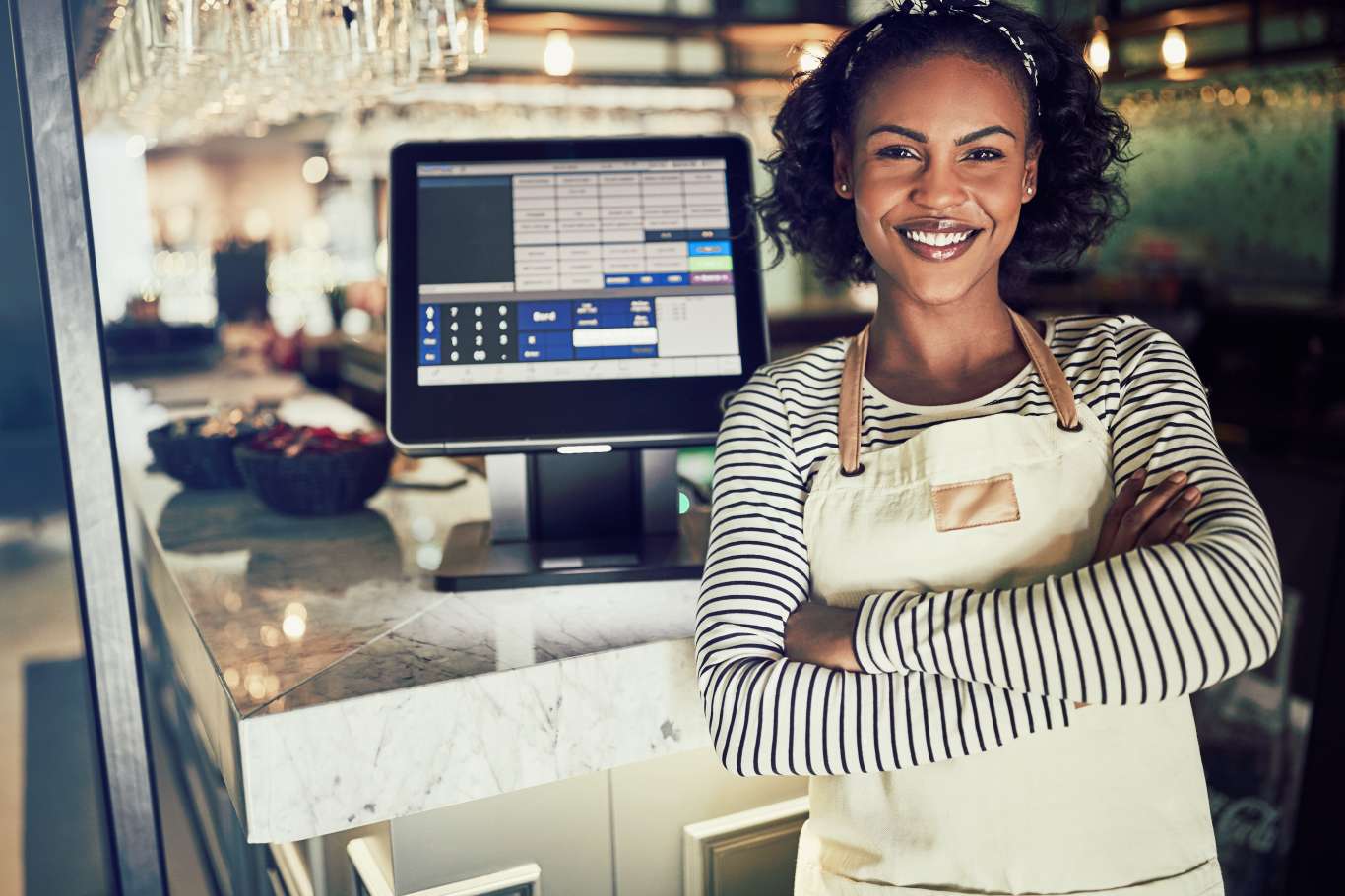 Young African waitress wearing an apron and smiling while standing by a point of sale terminal in a restaurant